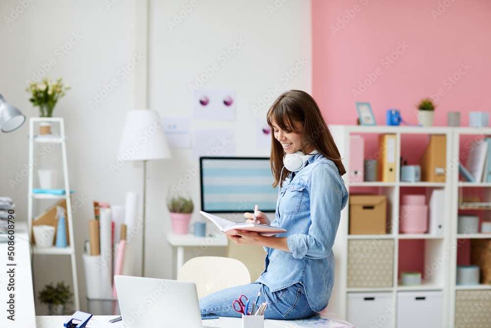 Cheerful creative young woman with brown hair sitting on table and analyzing notes in diary Cheerful creative young woman with brown hair sitting on table and analyzing notes in diary