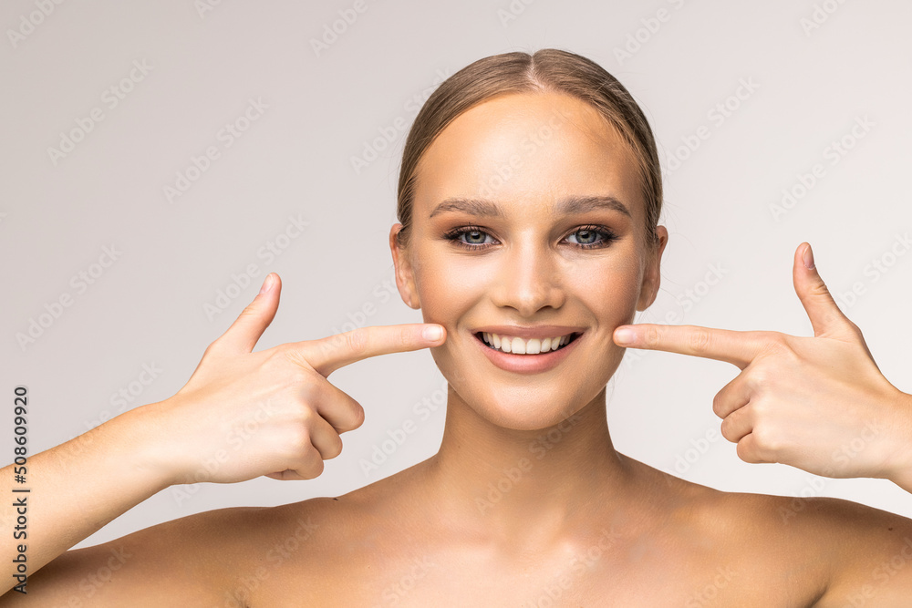 Young woman with perfect skin, bright spring pink make-up and naked shoulders on a white background. Pointing fingers at snow-white teeth.