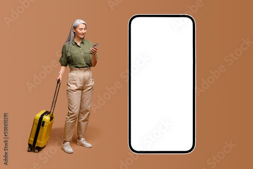 Travel app. Charming gray-haired mature modern woman stands with luggage near huge mobile phone with empty screen, senior Asian lady with yellow suitcase advertising app for booking and traveling