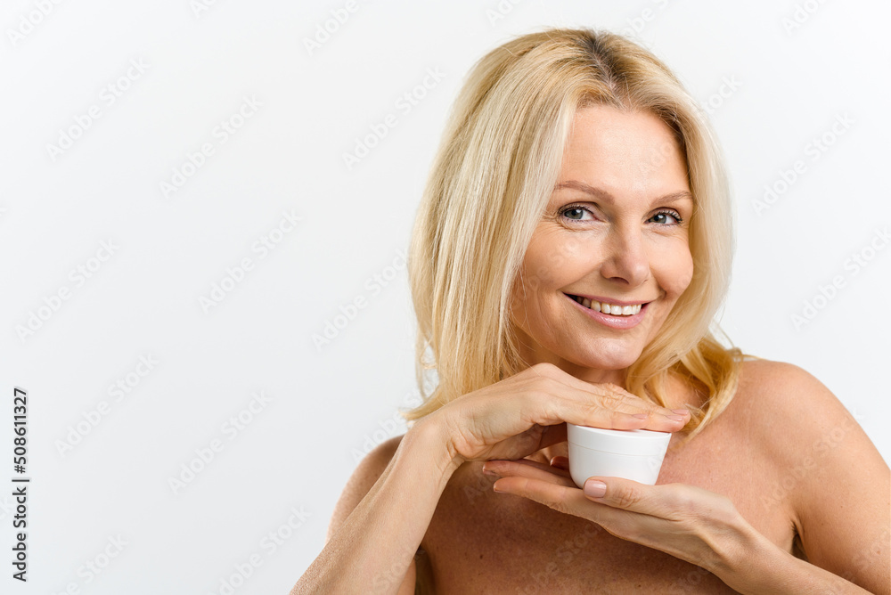Middle aged 50s woman holding jar with anti-aging moisturiser, preparing applying cream on the face, looking at the camera isolated on white. Skincare routine concept Middle aged 50s woman holding jar with anti-aging moisturiser, preparing applying cream on the face, looking at the camera isolated on white. Skincare routine concept