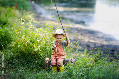 cute european boy in hat and overalls in summer with stick fishing rod fishes in the grass near the lake, plays fishing. Happy childhood in countryside