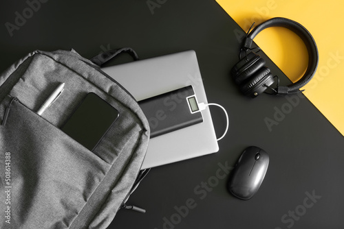 Gray textile urban backpack with laptop, external battery, phone, headphones, mouse and pen on black and yellow background. Travel, trip, study and lifestyle with gadgets. Recharging devices. photo