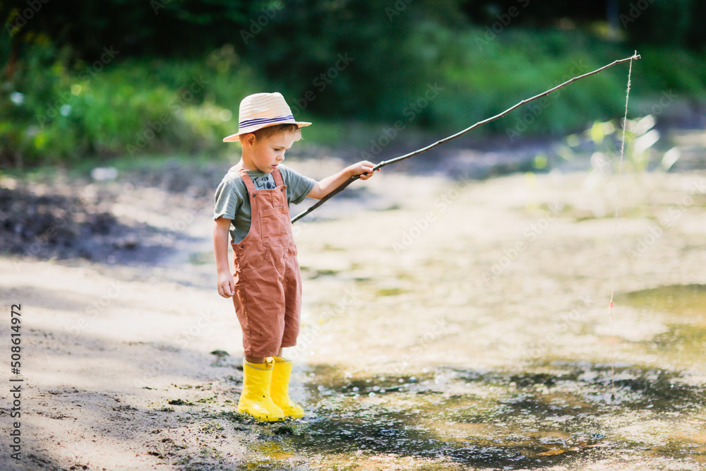 4-year-old child in wicker hat is fishing with stick, fishing rod and rope in grass by lake, boy play fishing. Happy childhood and holidays in the countryside, 4-year-old child in wicker hat is fishing with stick, fishing rod and rope in grass by lake, boy play fishing. Happy childhood and holidays in the countryside,