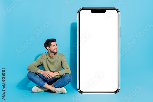 Full length portrait of satisfied person sit floor look empty space big phone isolated on blue color background