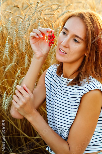 A young woman with a red currant berry in the form of an earring. A woman on the background of wheat ears at sunset. Portrait in nature.