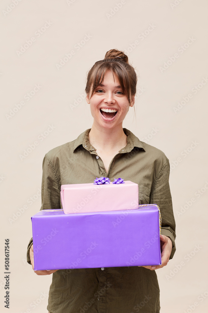 Thrilled beautiful young lady with bangs and hair bun standing against beige wall and holding stack of gift boxes Thrilled beautiful young lady with bangs and hair bun standing against beige wall and holding stack of gift boxes