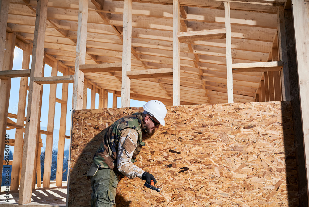 Carpenter hammering nail into OSB panel on the wall of future cottage