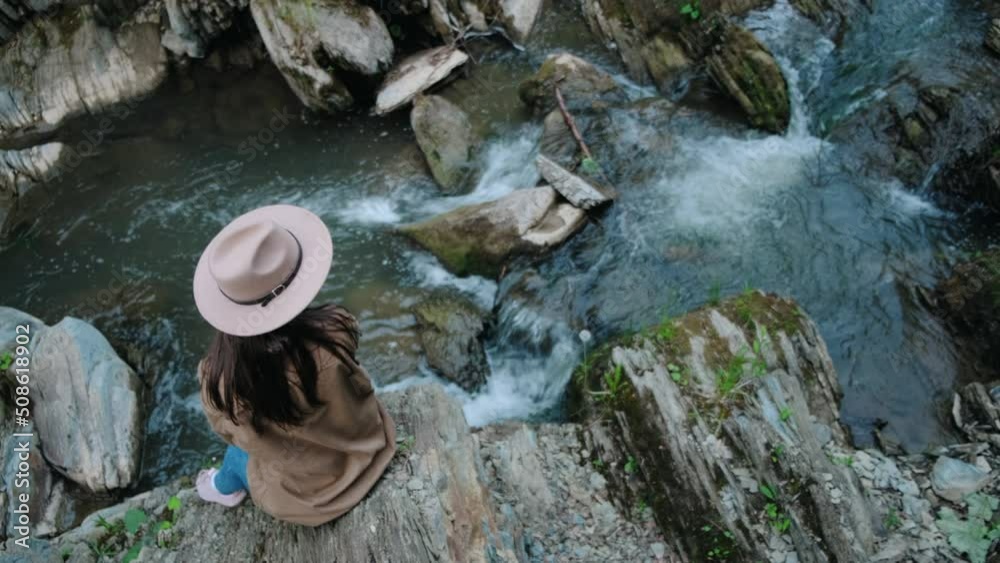 Simple and authentic young woman in hat looks around over amazing natural reserve in national park, mountain river. Hiking girl sitting on big stone. Explore outdoors, travel influencer blogger Simple and authentic young woman in hat looks around over amazing natural reserve in national park, mountain river. Hiking girl sitting on big stone. Explore outdoors, travel influencer blogger