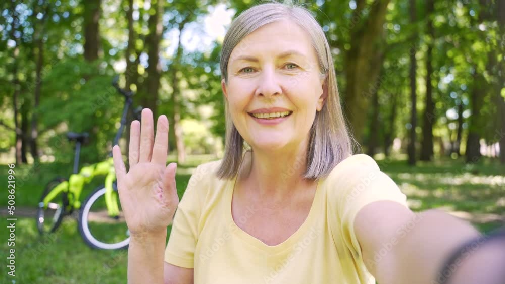 Cheerful senior woman standing in park, holding smartphone in arm, looking at camera talking and speaking while web calling, filming vlog smiling on a morning walk or vacation. View front camera phone