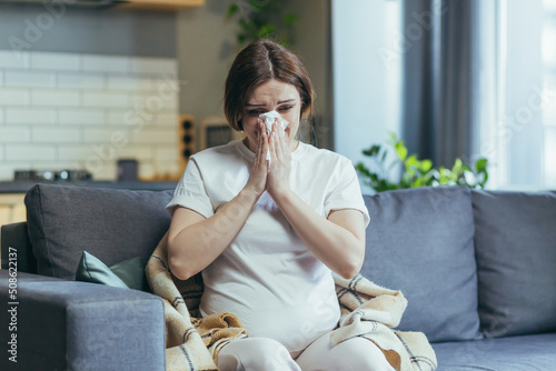 Sad pregnant woman at home sitting on the couch and crying, has a runny nose and allergies sneezes into a napkin photo