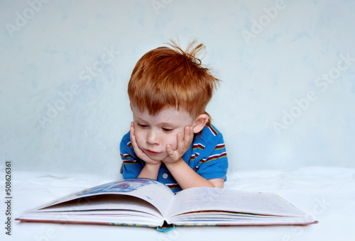 A cute little boy with red hair and a blue shirt is lying on the bed reading a book