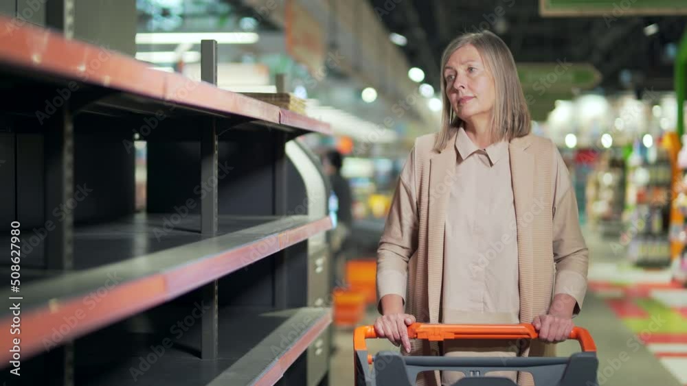 Senior woman shopper in supermarket, grocery store near empty shelves counters. Elder Mature female panic buying and stockpiling. groceries supplies with sold out products. Food supplies shortage