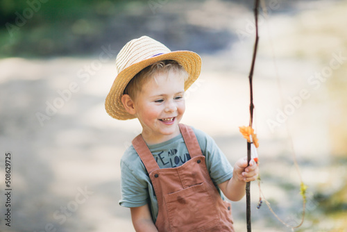 Preschooler boy in yellow rubber boots plays fishing, child fishes with stick near lake in countryside, games in nature and childhood without gadgets, summer holidays
