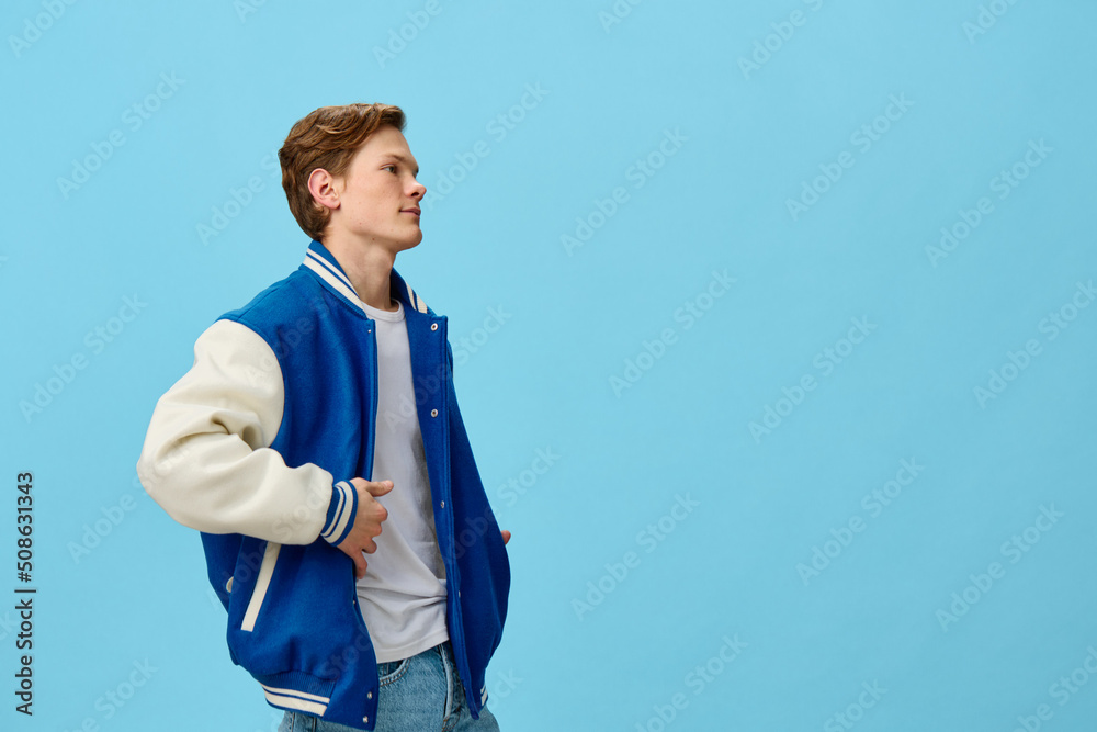 a handsome, fashionable guy in a blue bomber jacket is standing straightening his jacket on a plain background with empty space for text. studio photo