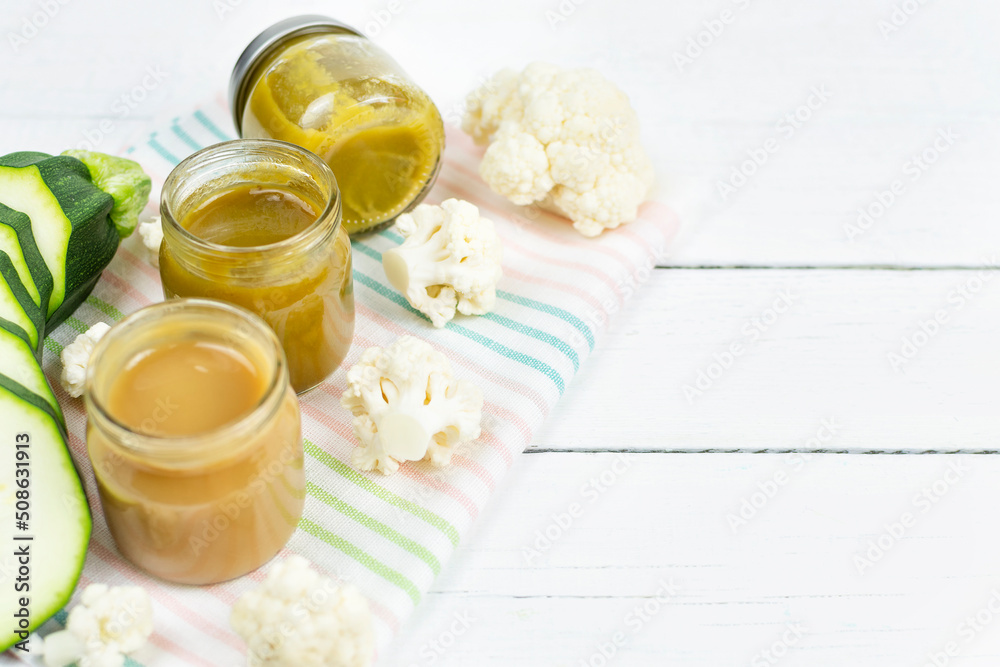 Glass jars with children's mashed cauliflower and zucchini puree on a white wooden background. Space for the text. Baby food. The first lure of the baby.