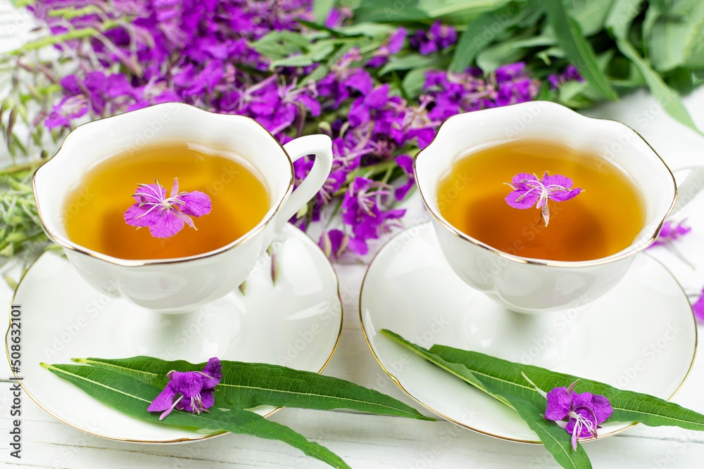 A drink (infusion, decoction) of ivan-tea in a white cup on a saucer on a wooden background. Grass and flowers of the plant kipreya, epilobium, ivan-grass. A drink (infusion, decoction) of ivan-tea in a white cup on a saucer on a wooden background. Grass and flowers of the plant kipreya, epilobium, ivan-grass.