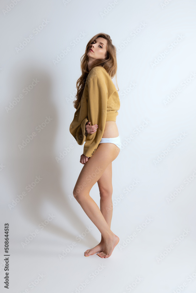 Full-length portrait of young stylish girl posing in yellow jacket and underwear isolated over grey studio background. Side view