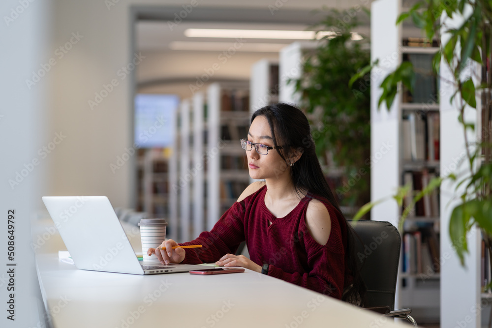 Focused asian female freelancer sitting in public library working remotely on laptop, concentrated korean girl studying online reading educational information in internet or preparing for lecture Focused asian female freelancer sitting in public library working remotely on laptop, concentrated korean girl studying online reading educational information in internet or preparing for lecture