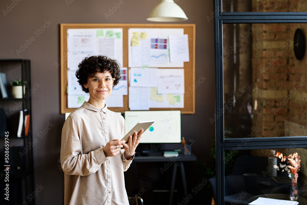 Portrait of young businesswoman with curly hair smiling at camera while working on digital tablet at office Portrait of young businesswoman with curly hair smiling at camera while working on digital tablet at office