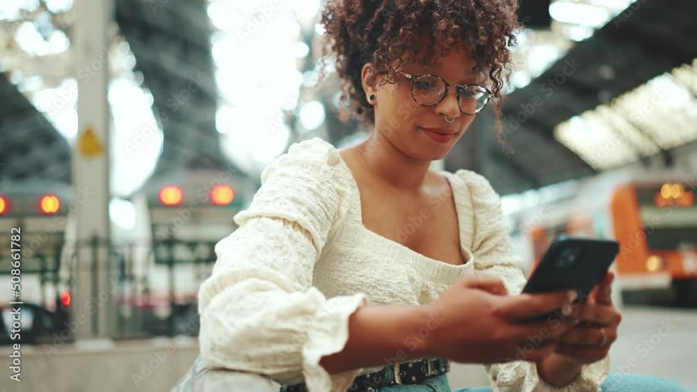Close-up of a young woman in glasses sits at the station, looks into a smartphone and smiles. Positive woman using mobile phone outdoors in urban background.