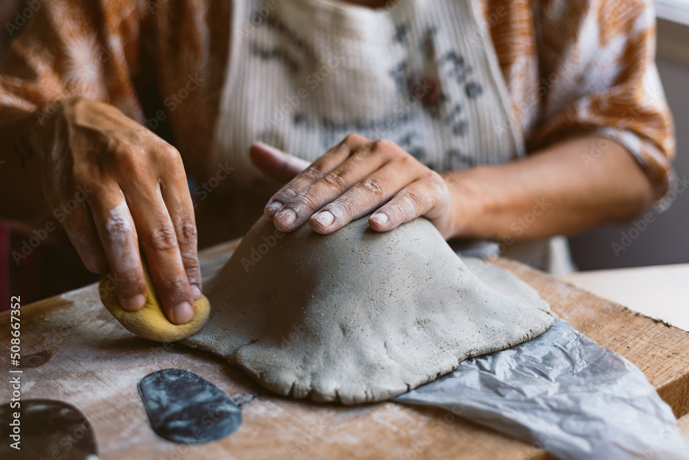 Close up of woman hands working clay making pottery at home. Concept of hobby and creativity at home. Kneading and moistening the clay.