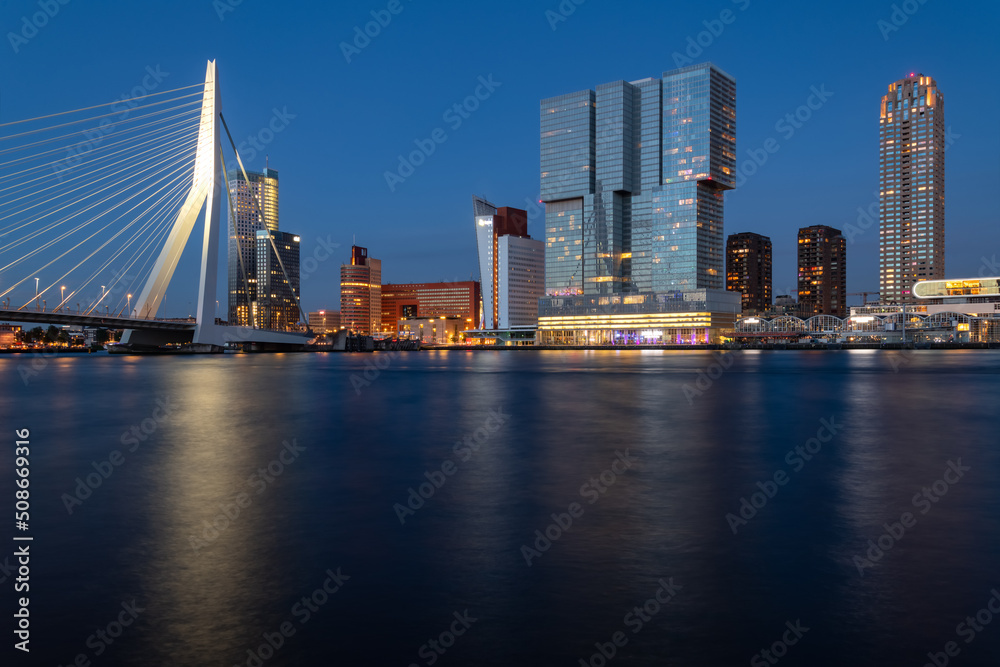 Rotterdam nighttime panorama with “Erasmus-Bridge“ over river Nieuwe Maas at evening blue hour in South Holland Netherlands. Waterfront with illuminated bridge and tall buildings on the waterfront.