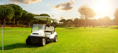 Golf cart in fairway of golf course with green grass field with cloudy sky and trees at sunset
