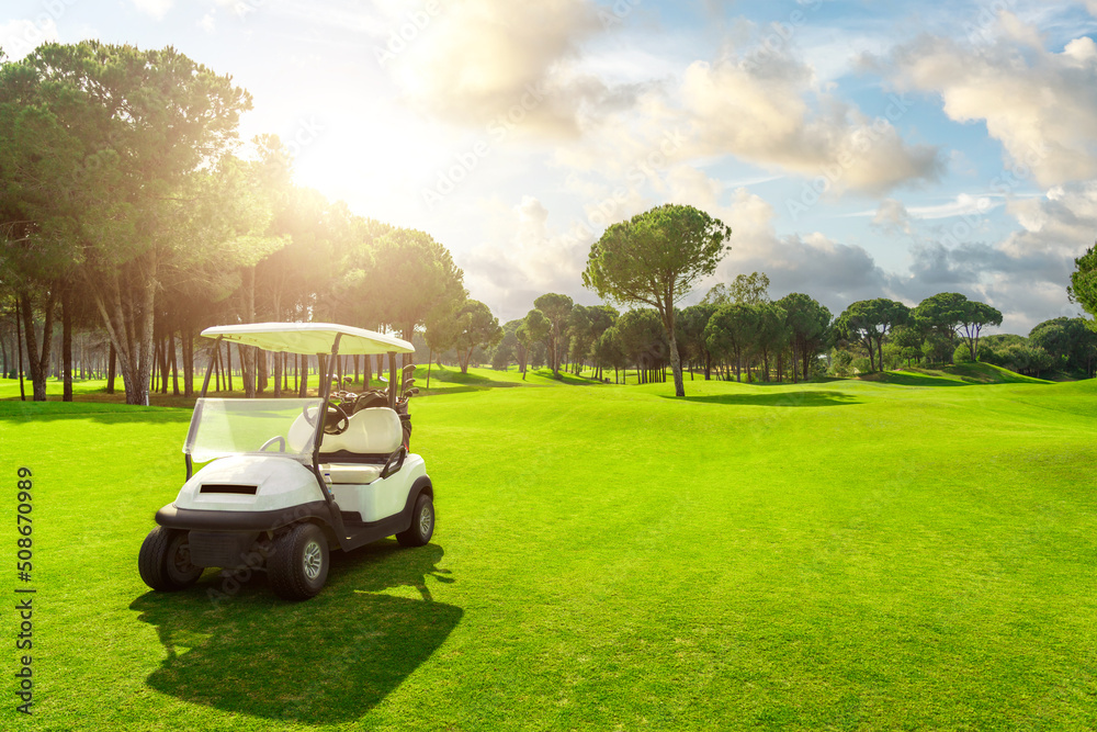 Golf cart in fairway of golf course with green grass field with cloudy sky and trees at sunset