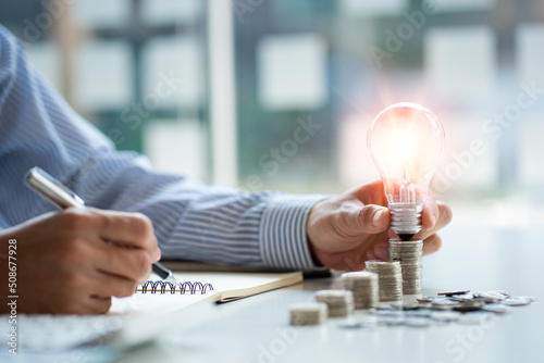 Businessman holding a light bulb on the left and a pen on the right. There are business report sheets and calculators on the table with stacks of coins and accounting whitening ideas.