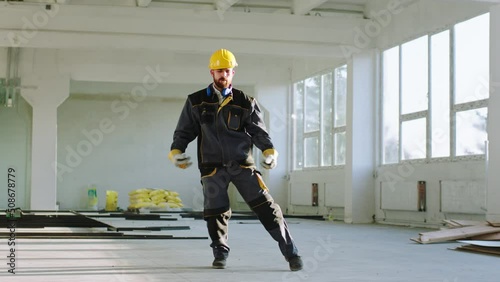 Attractive young engineer in a safety helmet and uniform dancing excited and happy at the end of the work day at modern construction building photo