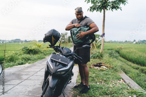 Young male tourist with backpack standing near black scooter with helmet parked at path way, casual dressed African American hipster guy traveling on vehicle moped during trip in Indonesia #508678910