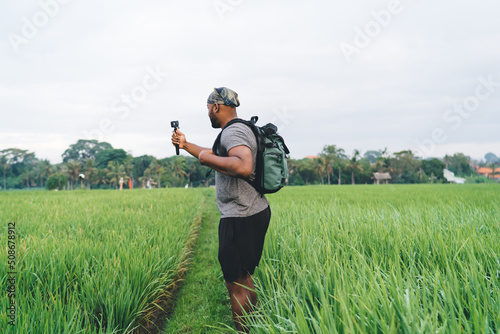 Side view of young male with backpack shooting influence content via waterproof modern camera exploring rice fields in Indonesia, millennial hipster guy creating video via digital technology © BullRun