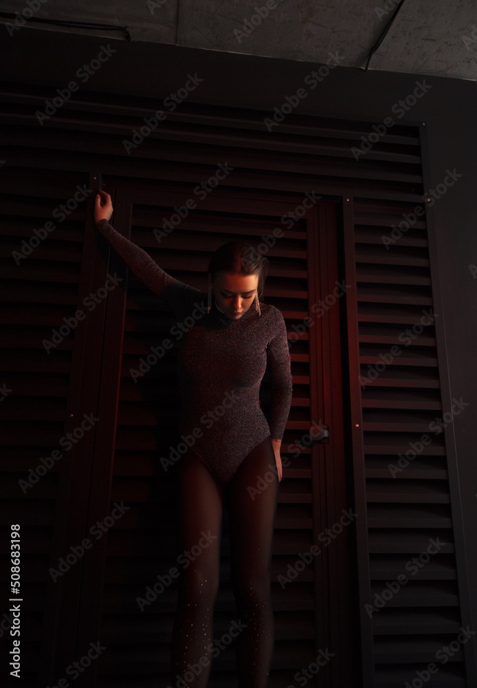 teen fashion. young brunette girl in a shiny silver bodysuit and boots is standing fashion with hand up and looking down near black iron wall background with red light. fashion concept, free space teen fashion. young brunette girl in a shiny silver bodysuit and boots is standing fashion with hand up and looking down near black iron wall background with red light. fashion concept, free space
