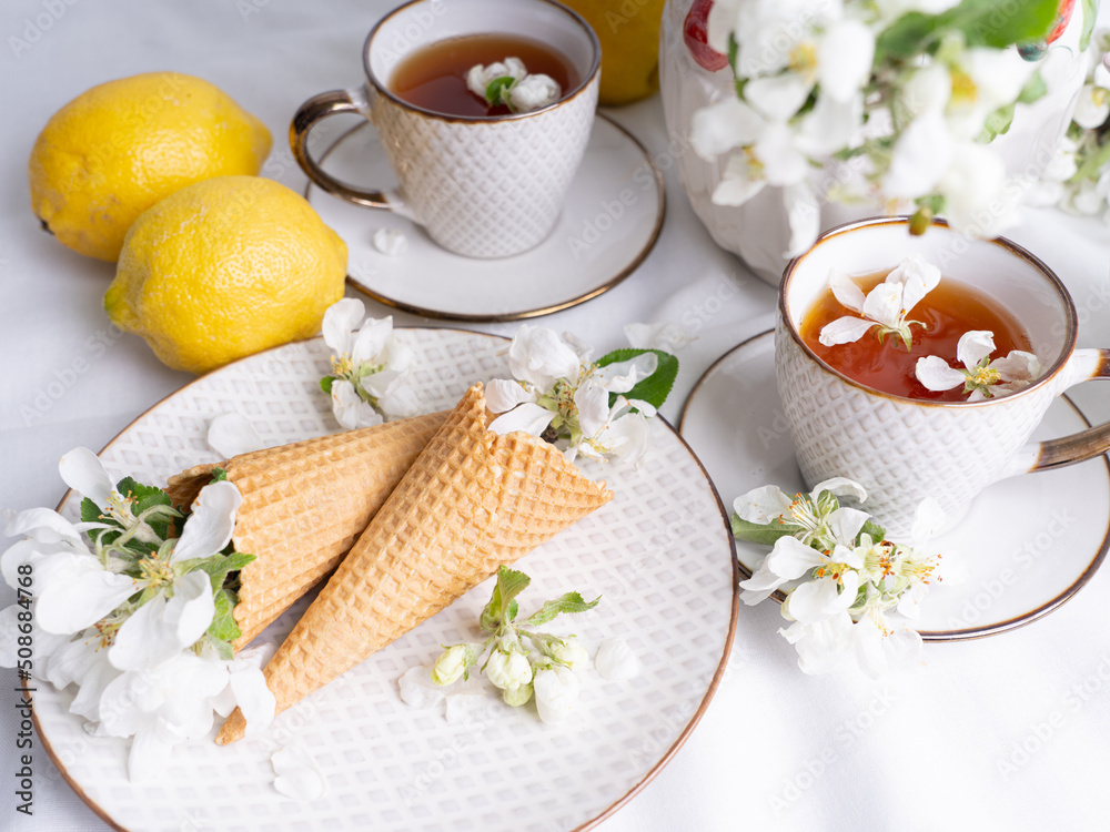 White tea set with tea in cups with yellow lemons and a blossoming apple branch..There are waffle cones on a saucer.
