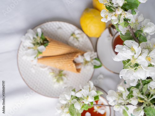 White tea set with tea in cups with yellow lemons and a blossoming apple branch.View from aboveThere are waffle cones on a saucer.