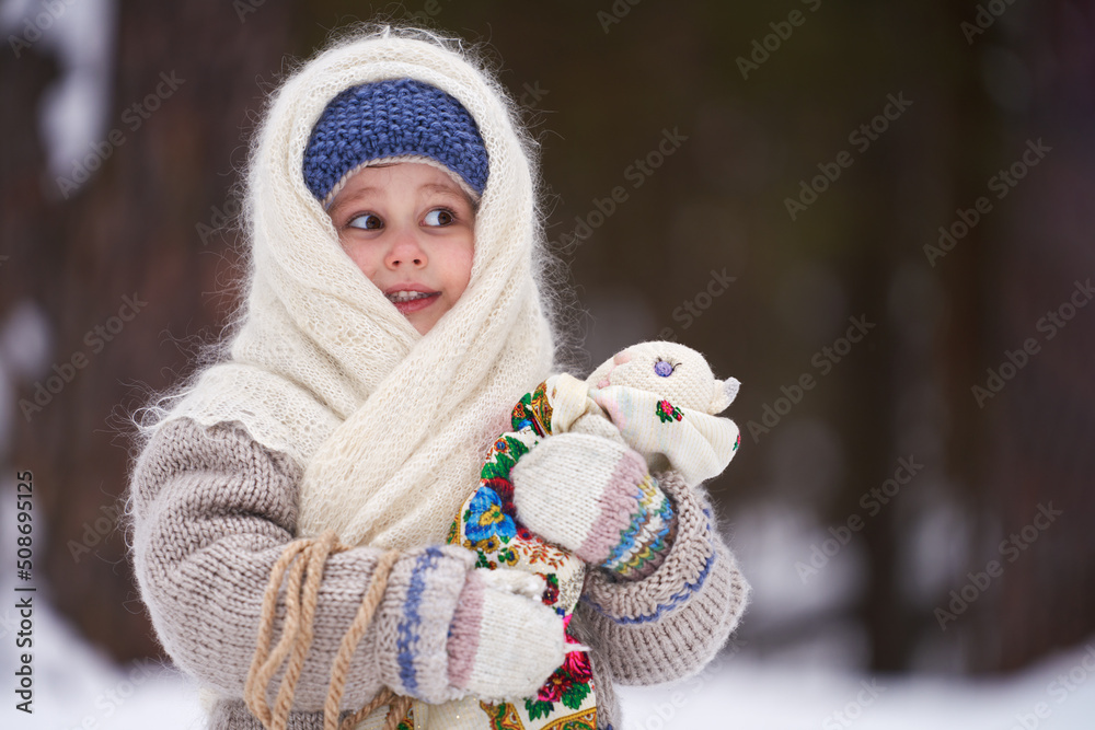 A little girl with a rag doll in her hands in a snowy winter forest. Rustic style. Vintage. Selective focus.