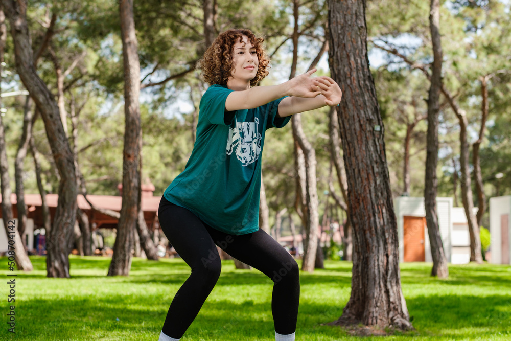 Young redhead woman wearing green t-shirt standing on city park, outdoor doing squats. Exercising her legs, outdoors sports concepts. Young redhead woman wearing green t-shirt standing on city park, outdoor doing squats. Exercising her legs, outdoors sports concepts.