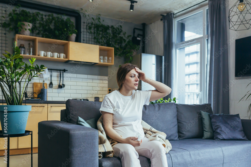 Obraz premium A young pregnant woman sits on the couch at home, has a runny nose and allergies, sneezes with a napkin