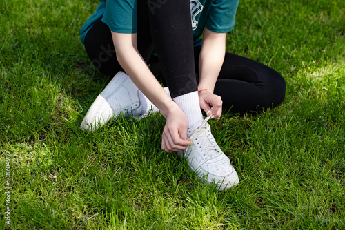 Close up view of young redhead woman wearing green tee standing on city park, outdoor tying lace running shoes getting ready for run. Jogging girl exercise motivation health and fitness.