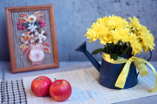 Still life of a bouquet of chrysanthemums in a decorative watering can, a picture in a frame and apples on a gray bokeh background.