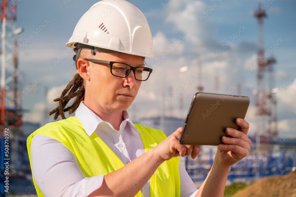 Portrait of woman inspector controls pipeline system for transporting light petroleum products using tablet on background of oil storage tanks. Portrait of woman inspector controls pipeline system for transporting light petroleum products using tablet on background of oil storage tanks.
