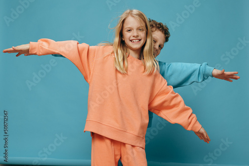 cute, beautiful, happy children a boy and a girl of school age stand on a blue background in bright plain clothes, facing the camera spreading their arms to the sides