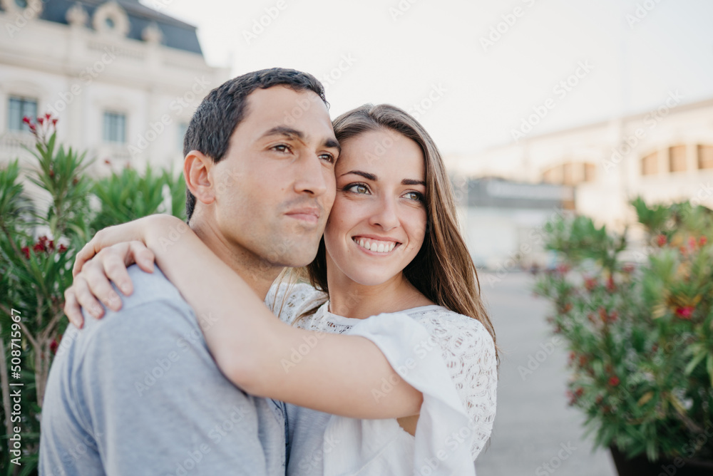 A smiling brunette woman is hugging her Hispanic boyfriend near the pier in Spain in the evening. A couple of tourists on a date in Valencia A smiling brunette woman is hugging her Hispanic boyfriend near the pier in Spain in the evening. A couple of tourists on a date in Valencia