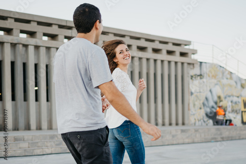 A smiling brunette girl and her Hispanic boyfriend are running on the pier in Spain in the evening. A couple of tourists on a date at the sunset in Valencia.