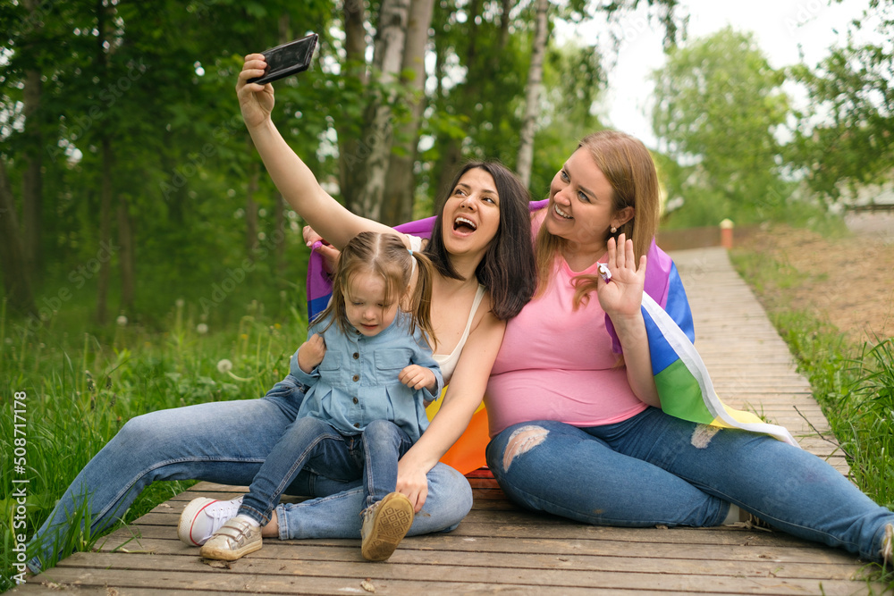 A happy married couple with a child take a selfie using their phone. Parents with an LGBT flag and a cute little girl are relaxing in nature in the park. LGBT parenting A happy married couple with a child take a selfie using their phone. Parents with an LGBT flag and a cute little girl are relaxing in nature in the park. LGBT parenting