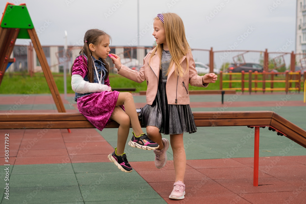 The child is caring for his girlfriend with a fractured arm and a plaster cast on her forearm. Girls have fun in yard and paint each other's lips with lipstick, sitting on playground bench