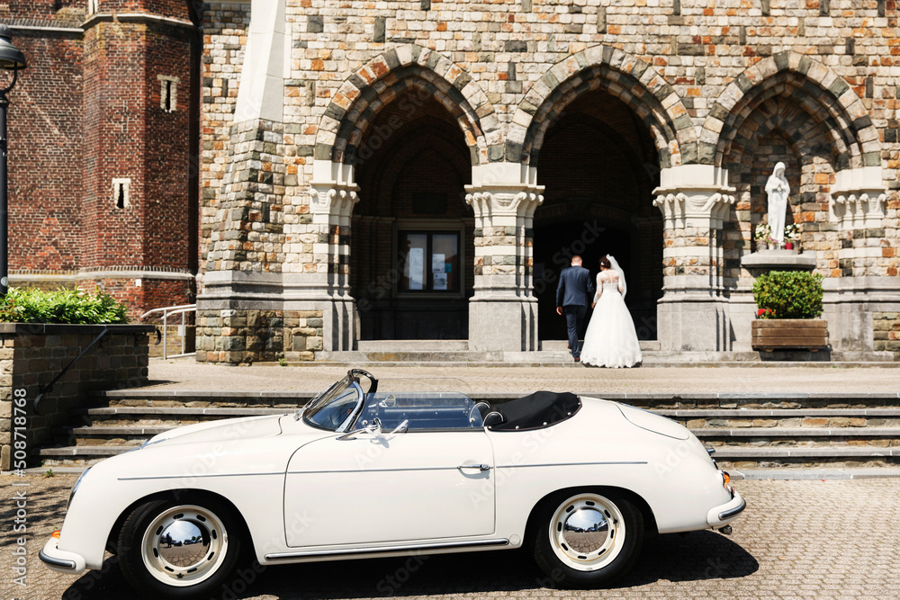 Newlyweds are walking up the stairs of the church and old white car