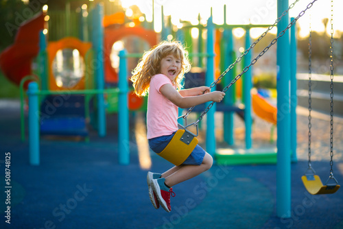 Excited child playing on outdoor playground. Kids play on kindergarten yard. Little boy having fun on a swing on the playground in public park on summer day. Happy child enjoy swinging.