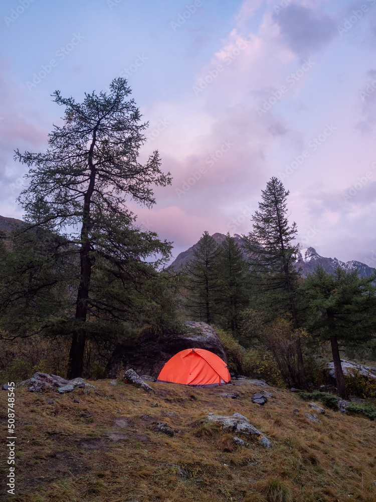 Twilight summer camp in the mountains. Amazing scenery of twilight in mountains with tent on pass in violet light. Tent near rocky hill in lilac light. Purple sunset light.