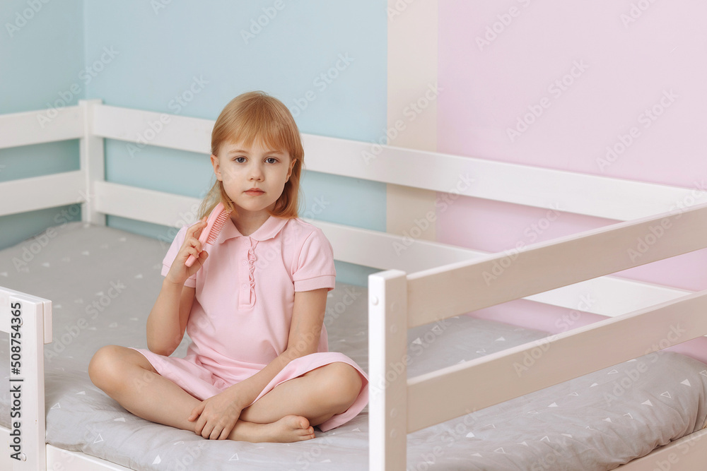 A little preschool girl in a pink dress sits on the bed combing her hair. A little preschool girl in a pink dress sits on the bed combing her hair.